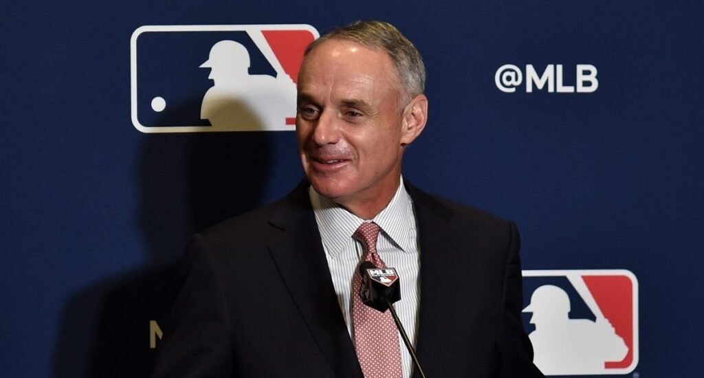 Rob Manfred speaking at an MLB press conference, MLB logo